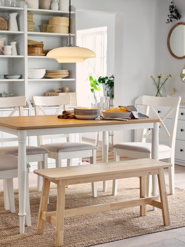 Table en chêne/blanc, quatre chaises blanches, banc en bouleau et tapis en jute. Deux grands meubles à l'arrière-plan.