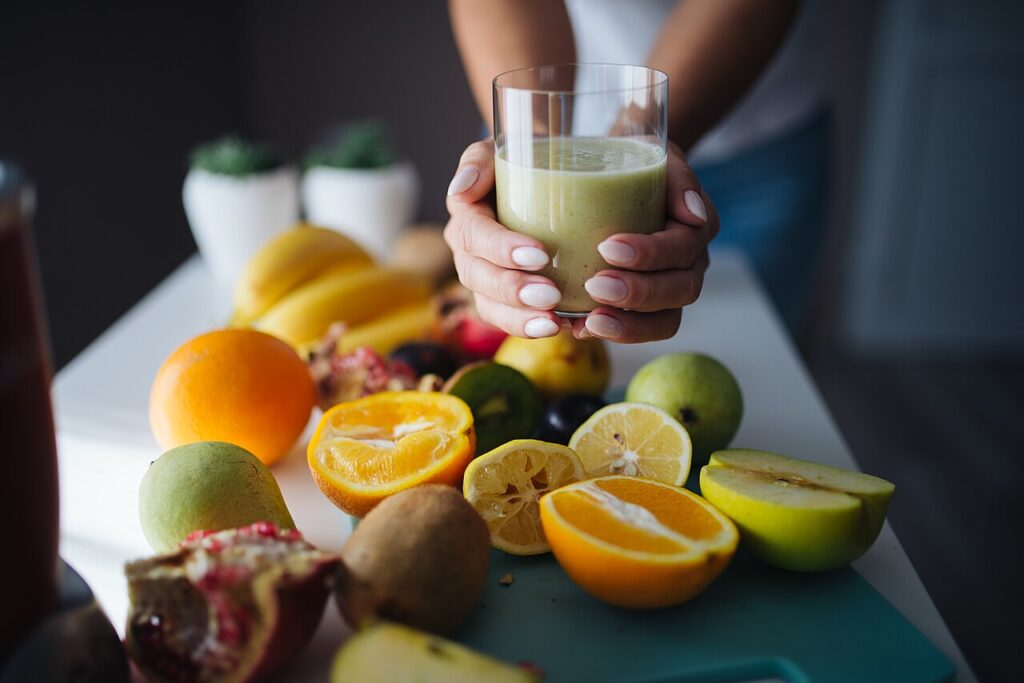 15 Porte - Bright kitchen filled with fresh fruits like oranges, lemons, and pomegranates. A person holds a green smoothie above a colorful fruit arra